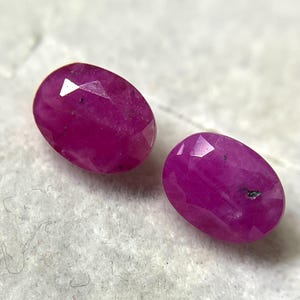 Peut inclure: Deux rubis ovales à facettes d'une couleur magenta-rose profond. Chaque pierre précieuse présente une surface polie avec des inclusions internes visibles et des facettes géométriques qui reflètent la lumière sur leurs surfaces vibrantes.