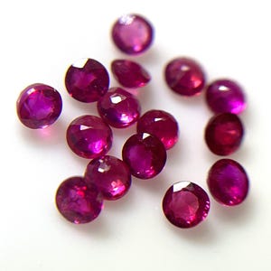 May include: A collection of small, round, faceted loose gemstones in a vibrant magenta-red colour. Each stone features a brilliant cut with multiple reflective surfaces that catch the light, creating a sparkling appearance across the group of gems.