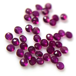 Puede incluir: Una colección de pequeñas piedras preciosas redondas y facetadas de color magenta rosado intenso. Cada piedra presenta múltiples superficies planas que captan la luz, creando un efecto brillante en el grupo de gemas circulares individuales.