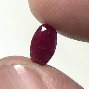 May include: A deep red, oval-shaped faceted gemstone with a polished surface. The stone exhibits a rich, saturated crimson hue and clear geometric facets that reflect light, showcasing its translucent quality and precise cut in a vertical orientation.
