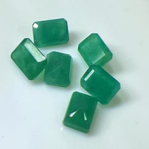 May include: Emerald-cut green gemstones. The gemstones are a vibrant green color and have a rectangular shape with cut corners. The facets catch the light, creating a sparkling effect. The gemstones are scattered on a white surface.