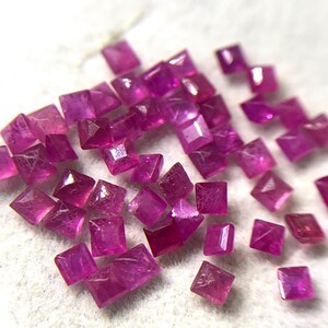 May include: A collection of small, square-cut, vibrant magenta-pink gemstones. Each stone features a faceted surface that reflects light, creating a sparkling appearance. The stones are arranged in a loose cluster, showcasing their uniform shape.