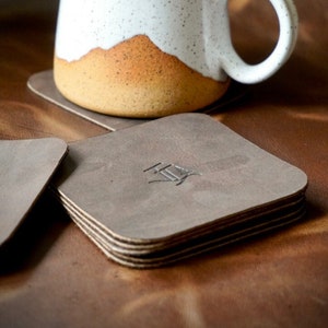 Puede incluir: Una taza de cerámica blanca con motas marrones y un borde marrón se asienta sobre una pila de cuatro posavasos de cuero gris con la palabra "HEY" grabada en el posavasos superior.
