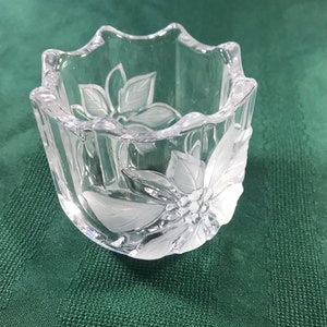 May include: A clear glass votive candle holder with a frosted floral design. The holder has a scalloped rim and a square shape. The frosted design features a flower and leaves. The candle holder is sitting on a green surface.