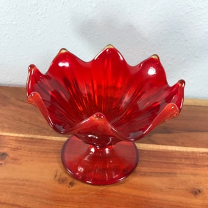 May include: A vibrant red glass bowl with a scalloped rim and a pedestal base. The bowl's design resembles a flower, with each petal-like curve extending outwards. The glass has a glossy finish, reflecting light and adding to its visual appeal. The bowl is displayed on a wooden surface.
