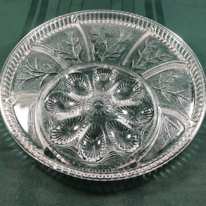 May include: A clear glass serving dish with six individual compartments for serving oysters. The dish has a decorative etched design of leaves and branches around the edge.