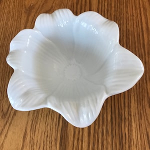 May include: A white ceramic flower-shaped dish with a smooth, glossy finish. The dish has five petals and a rounded center.