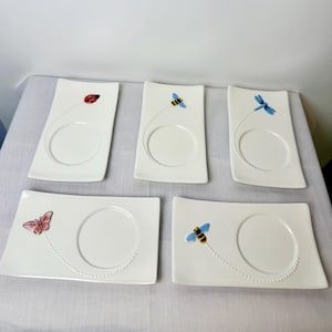 May include: A set of five rectangular white ceramic plates, each featuring a circular indentation and a small raised insect figure. The insects include a ladybug, a bee, a dragonfly, a butterfly, and another bee, each with a dotted trail.