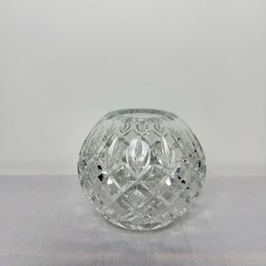 May include: A clear, spherical crystal vase featuring a detailed diamond-cut pattern across its entire surface. The glass object has a circular opening at the top and a rounded body, reflecting light through its intricate, faceted geometric design.