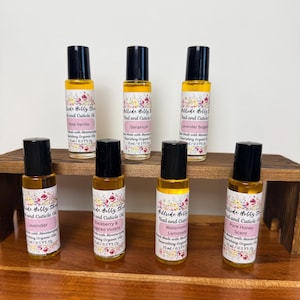 May include: Seven roll-on bottles of nail and cuticle oil are displayed on a wooden shelf. Each bottle has a black cap and a label with floral designs and text indicating different scents like Rose Vanilla, Geranium, and Lavender. The bottles are 15 ml / 0.5 fl oz.