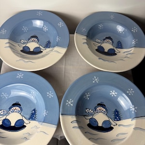May include: Four ceramic plates with a winter theme. Each plate features a blue rim with white snowflakes, a snowman design, and a white base. The snowman is wearing a hat and scarf, and there is a small pine tree.