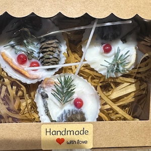 以下が含まれることがあります： 白い貝殻型のキャンドルが3つ入った箱。各キャンドルには、松葉、ベリー、松ぼっくりなどの自然な要素が飾られています。箱には「Handmade with love」と書かれたステッカーが付いています。