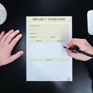 Puede incluir: Una hoja de papel blanca con un encabezado amarillo que dice "PROJECT OVERVIEW". El papel está dividido en secciones para la descripción del proyecto, los objetivos, la fecha de inicio, la fecha límite, los hitos, los recursos y una lista de tareas pendientes. Una mano está escribiendo en el papel con un bolígrafo negro.