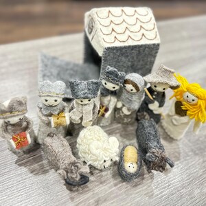 Peut inclure: Une crèche de Noël en feutre avec une église grise, un mouton blanc et des figurines des trois rois mages, Marie, Joseph et un ange. Les figurines sont toutes habillées en feutre gris et blanc.