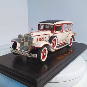 May include: A cream-colored vintage car model with red accents and a black roof, mounted on a dark wooden base. The car features white-walled tires and red wheels. The model is a detailed replica of a classic automobile.