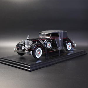 May include: A black scale model vintage car featuring a chrome grille, white-walled tires with red center caps, and a spare tire mounted on the side. The vehicle has a black convertible top and thin red pinstriping along the body and fenders.