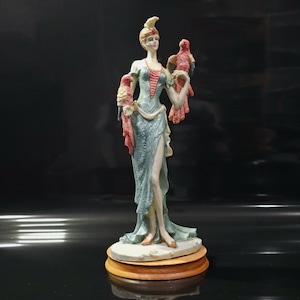 Puede incluir: Figura decorativa de una mujer con un vestido fluido color verde azulado, sosteniendo dos loros rojos y beige. La estatua tiene una base de color crema y un soporte de madera. El vestido de la mujer tiene una abertura alta.
