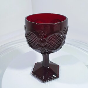 Op de afbeelding: Een robijnrode kelk met een ruitpatroon. Het glas heeft een zeshoekige basis en een steel die verbinding maakt met de kom. De kom heeft een decoratief patroon van cirkels en ruiten. De kelk is gemaakt van glas.