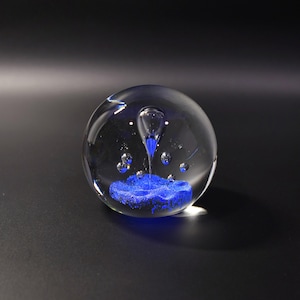 May include: A clear glass paperweight shaped as a sphere containing a textured blue base, a central blue teardrop-shaped inclusion, and several small suspended air bubbles distributed throughout the transparent glass body.