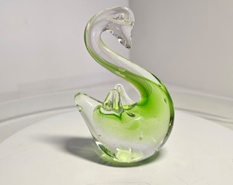 May include: A clear glass swan figurine with green accents. The swan has a long, curved neck and a detailed beak. The body of the swan is filled with a vibrant green color, and the glass has a smooth, glossy finish. The figurine is a decorative item.