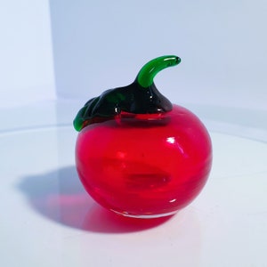 Puede incluir: Una figura de manzana de cristal rojo vibrante, con un tallo y hojas verdes. La fruta brillante y transparente es un objeto decorativo. La manzana tiene una forma clásica, con una forma lisa y redondeada.