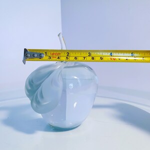 May include: A frosted glass apple sculpture with a stem, measuring approximately 10 cm in length. The apple has a light blue hue and a textured leaf design on one side. The sculpture is on a white surface.
