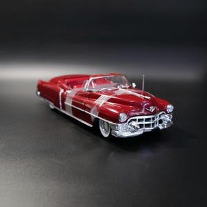 May include: A die-cast scale model of a vintage deep red convertible car featuring a prominent chrome front grille, circular headlights, white-wall tires, and a detailed interior. The vehicle includes a silver antenna and classic body styling.