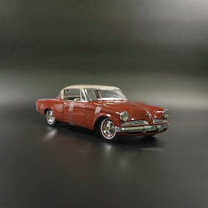 May include: A scale model of a vintage two-door coupe motor car featuring a deep terracotta red body and a cream-coloured roof. The vehicle has chrome bumpers, circular headlights, white-wall tyres, and a detailed interior visible through the windows.