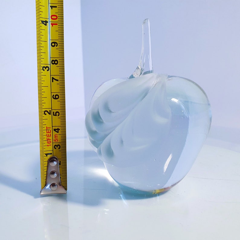 May include: A clear glass apple-shaped ornament with a stem. The apple is approximately 10 cm tall. The glass has a frosted, swirled design in shades of white and light blue. The object is sitting on a reflective surface.
