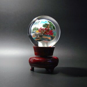 Può includere: Una sfera di vetro trasparente con un paesaggio dipinto all'interno, posta su un supporto in legno rosso scuro. Il paesaggio presenta alberi, montagne e acqua nelle tonalità del verde, rosso e blu. Il supporto ha una base rotonda e tre piedini.