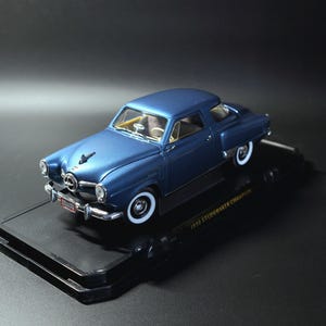 May include: A blue 1950 Studebaker Champion model car with white-walled tires. The miniature car has a detailed chrome grill and trim. The model sits on a black display base.