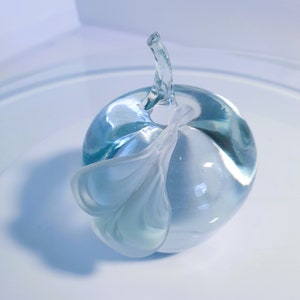 May include: A clear glass apple sculpture with a stem and a leaf-like design. The apple is a light blue colour and has a smooth, rounded shape. The leaf is white and has a delicate, flowing form. The sculpture is likely a decorative item.
