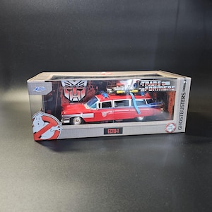Può includere: Un veicolo Ecto-1 rosso e blu pressofuso del franchise Ghostbusters, confezionato in una scatola con il marchio Transformers. La scatola presenta il logo Ghostbusters e il testo "ECTO-1".