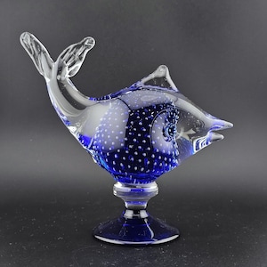 May include: A clear and cobalt blue glass fish sculpture. The fish has a detailed design with bubbles in the body and fins. It is mounted on a pedestal base. The sculpture is a decorative art piece.