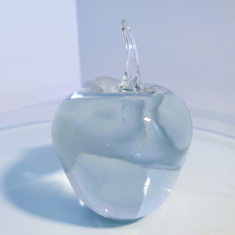 May include: A clear glass apple sculpture with a curved stem. The apple is smooth and rounded, with a slight blue tint. The stem is elongated and curved, adding a touch of elegance to the design. The sculpture is displayed on a white surface.