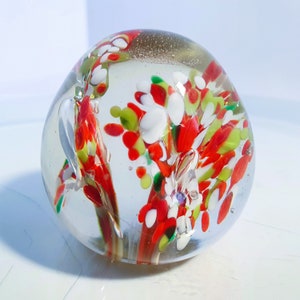 May include: A clear glass paperweight with colourful internal designs. The paperweight features red, white, and green abstract floral patterns. The glass is smooth and rounded, with a few small bubbles visible within the glass.