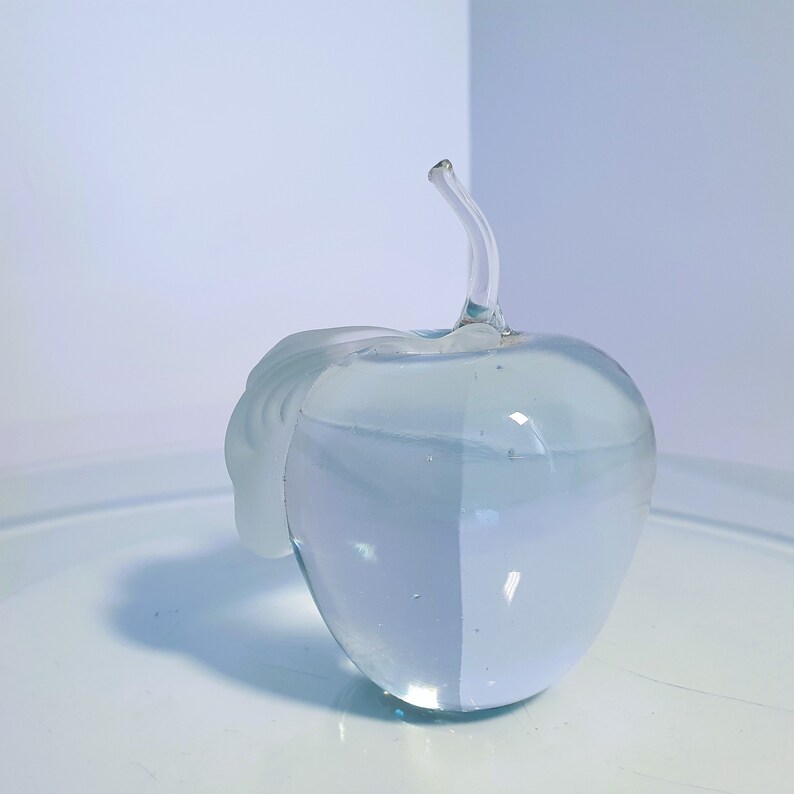 May include: A clear glass apple sculpture with a curved stem and a frosted leaf detail. The apple is displayed on a white surface, with a light blue background. The sculpture is a decorative item.