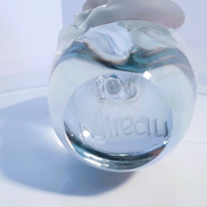 May include: A clear glass decorative object with a rounded base and a frosted, petal-like top. The word "Aireau" is etched into the base. The object is transparent, allowing light to pass through, creating shadows.
