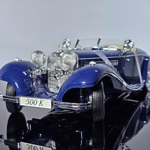 Puede incluir: Modelo detallado de un coche descapotable azul clásico con detalles cromados. El coche presenta faros grandes, una parrilla prominente y el texto "500 K" en la parte delantera. El coche se exhibe sobre una superficie reflectante.