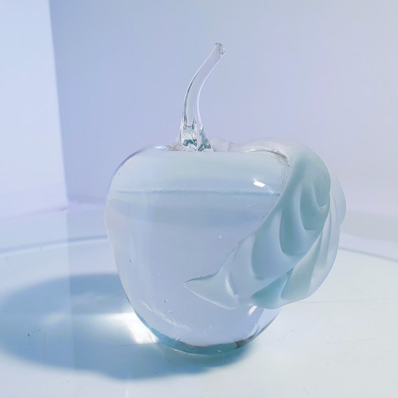 May include: A clear glass apple sculpture with a curved stem and a leaf-like design on the side. The apple is transparent, allowing light to pass through, and sits on a reflective surface. The sculpture is a decorative piece.