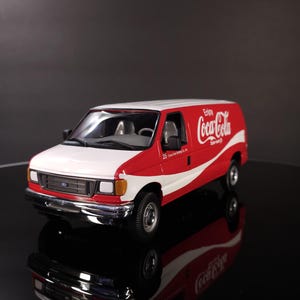 May include: A red and white Coca-Cola delivery van model. The van has the Coca-Cola logo on the side and the text "Enjoy Coca-Cola". The model is a Ford van with a white roof and a red body.