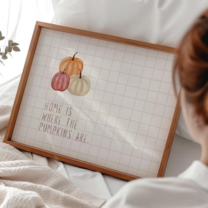 Pode incluir: Uma impressão emoldurada em madeira com três abóboras em aquarela em tons de laranja, vermelho e creme. A impressão tem um fundo quadriculado e o texto "HOME IS WHERE THE PUMPKINS ARE."