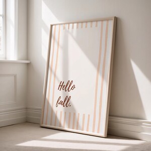 May include: A framed art print with a beige background and vertical peach stripes. The words "Hello fall." are written in a brown script font. The frame is a light wood color, and the print is leaning against a white wall.