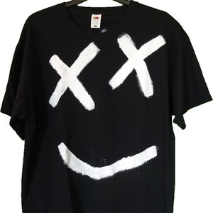 Op de afbeelding: Zwart t-shirt met een wit smiley-gezicht ontwerp. Het ontwerp heeft twee witte 'X'-vormen voor ogen en een gebogen witte lijn voor een mond. Het shirt hangt aan een zwarte hanger.