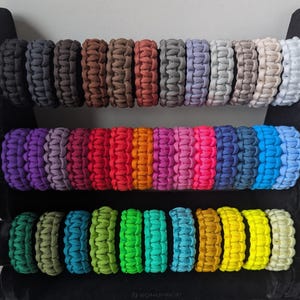 Könnte beinhalten: Eine Auswahl von 30 bunten Paracord-Armbändern, jedes mit einer einzigartigen Farbkombination. Die Armbänder sind in drei Reihen zu je zehn angeordnet, wobei jede Reihe ein anderes Farbschema aufweist. Die Armbänder sind aus strapazierfähigem Paracord gefertigt und eignen sich perfekt für Outdoor-Aktivitäten, Überlebenssituationen oder den täglichen Gebrauch.
