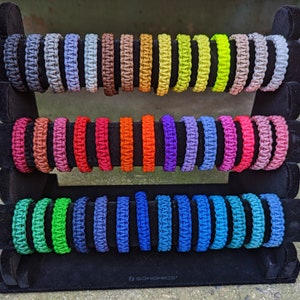Op de afbeelding: Een display met 24 kleurrijke paracord armbanden, elk met een uniek gevlochten patroon. De armbanden zijn gerangschikt in drie rijen op een zwarte standaard met de tekst "#SONGMICS" aan de onderkant.