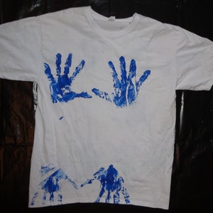 Op de afbeelding: Wit T-shirt met blauwe handafdrukken. Twee handafdrukken zijn op de borst, en meer afdrukken bij de zoom. Het shirt is een casual, korte mouwen stijl.