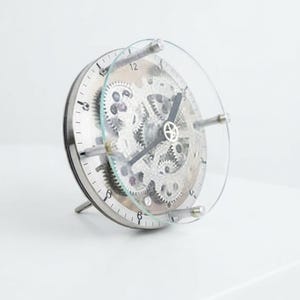 May include: A circular table clock with a transparent glass front and a silver-toned metal frame. The clock face reveals an intricate internal mechanism featuring exposed silver gears and cogs, with black hour and minute hands centred.