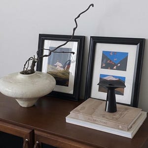 May include: A low, wide, off-white ceramic vase with a textured finish containing a thin, dark, twisted branch. Next to it, a small black trumpet-shaped vase sits on two stacked books titled All Directions, flanked by two framed art prints.