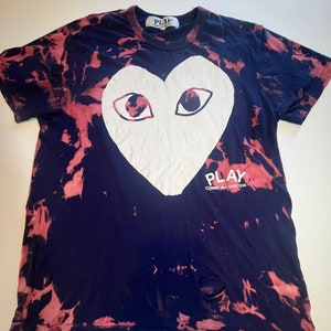Puede incluir: Una camiseta azul marino con un gráfico de corazón y ojos blancos. La camiseta tiene un patrón tie-dye en tonos de rosa y azul. El texto "PLAY COMME des GARCONS" está impreso en la parte inferior del corazón.
